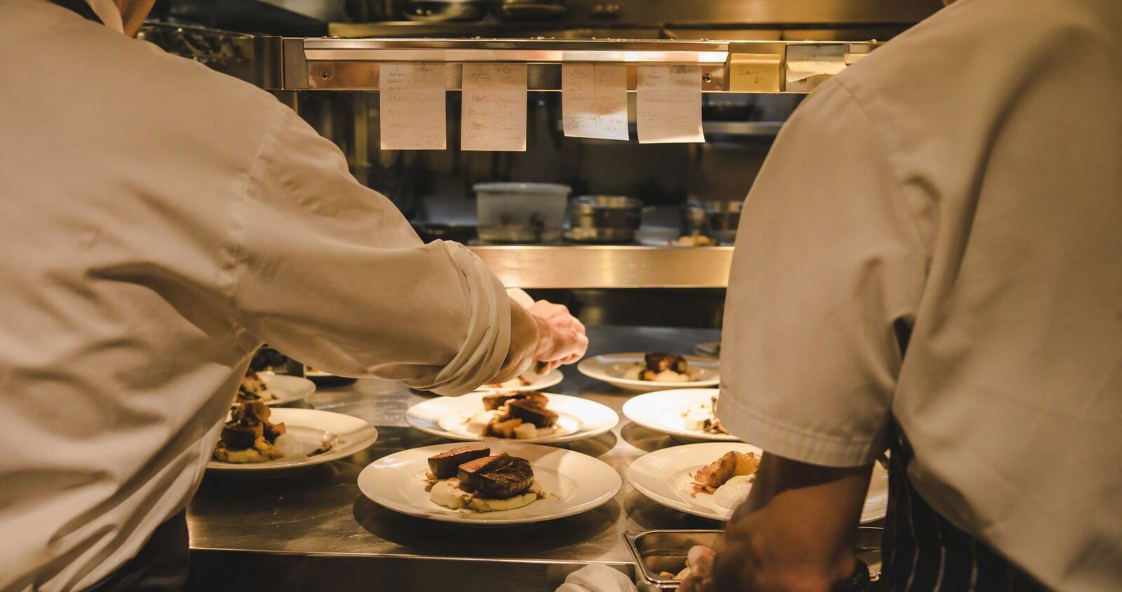 Chefs plating courses at the pass, tickets on the rail, warm kitchen light